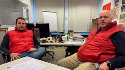 Zwei Männer sitzen in einem Büro. Sie haben rote Westen an und schauen in die Kamera