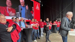 Das Foto zeigt rot gekleidete Menschen, die hinter einem Redner auf einer Bühne stehen. 