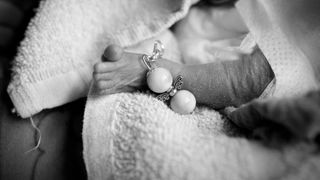 Das Bild ist schwarz weiß. Darauf ein ganz kleiner Babyfuß, der eine Fußkette aus zwei Perlen trägt, die einen Engel darstellen sollen. Der Fuß gehört zu einem verstorbenen Baby, das ist einem hellen Handtuch liegt.