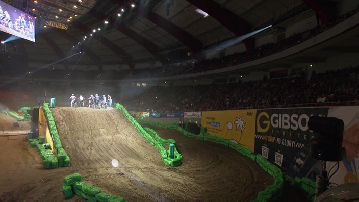 6 Motocross-Fahrer auf der Piste im Stadion, im Hintergrund sieht man das Publikum