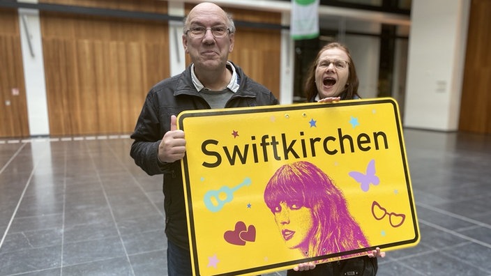 Ein Mann und eine Frau halten ein gelbes "Swiftkirchen"-Schild. Beide lachen und freuen sich. 