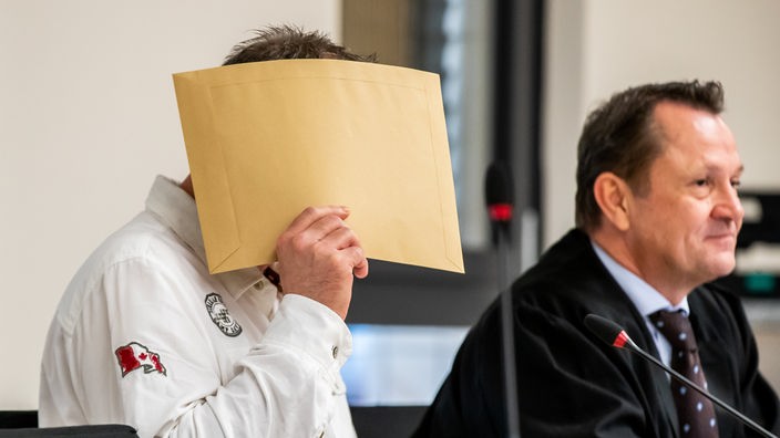 Ein Mann hält sich einen Umschlag vor das Gesicht, neben ihm sitzt ein Mann in einer Robe.
