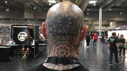 Ein Mann mit komplett tätowiertem Kopf von hinten, im Hintergrund Messestände
