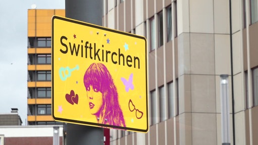 Ein gelbes Schild mit der Aufschrift "Swiftkirchen" und einem pinken Konterfei der Sängerin Taylor Swift hängt in Gelsenkirchen