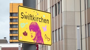 Ein gelbes Schild mit der Aufschrift "Swiftkirchen" und einem pinken Konterfei der Sängerin Taylor Swift hängt in Gelsenkirchen