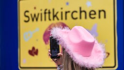 Das Ortsschild "Swiftkirchen", davor eine Frau mit pinkem Hut auf dem Kopf