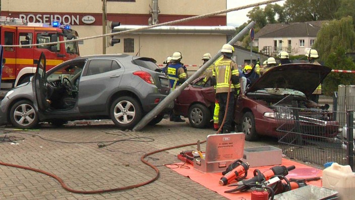 Zwei zerstörte Autos an einer krummen Laterne, dahinter Rettungskräfte mit Werkzeug