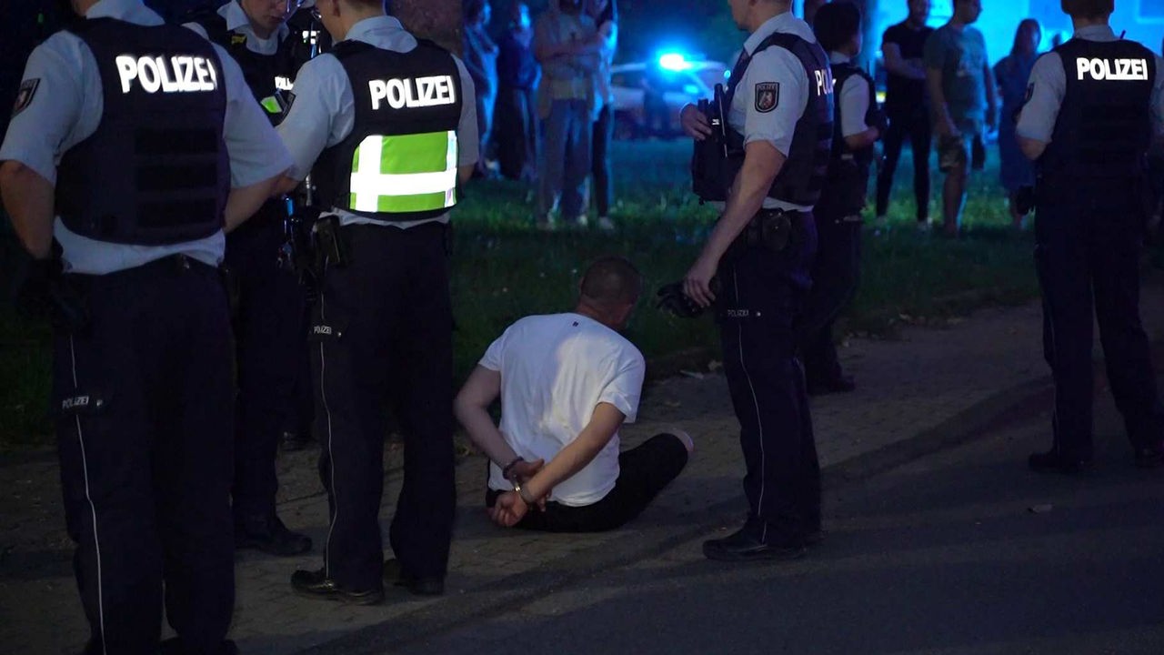 Auf dem Foto ist ein Mann, der in Handschellen auf dem Boden sitzt. Um ihn herum stehen Polizisten.