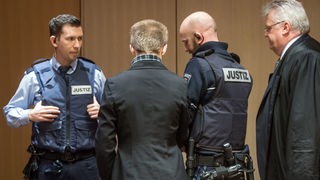 Marcel H. steht in schwarzer Jacke im Gerichtssaal mit dem Rücken zur Kamera, neben Wachtmeistern und seinem Anwalt