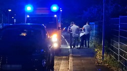 Drei Personen halten eine weitere Person in gebückter Haltung fest. Daneben steht ein Krankenwagen. 