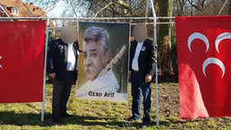 Zwei Männer stehen vor einem Portraitfoto von Ozan Arif