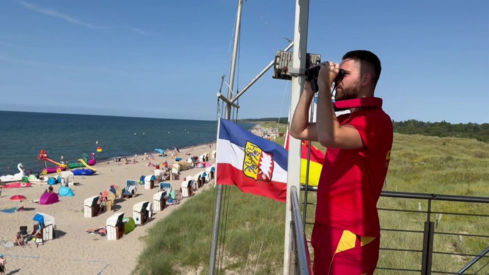 Das Foto zeigt den Rettungsschwimmer in roter DLRG-Kleidung auf einem Aussichtsposten am Strand mit Fernglas