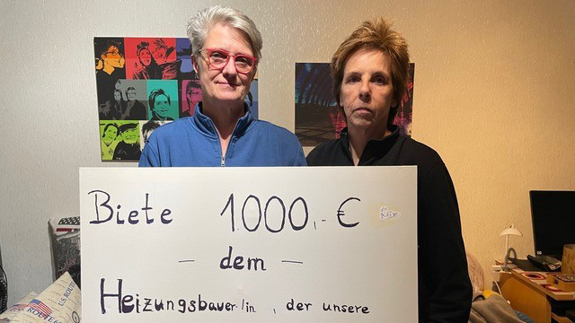 Ein paar steht mit einem Schild in ihrer Wohnung, auf diesem steht: Biete 1000€ dem Heizungsbaer/in, der unsere schlafraubende Heizungsgeräusche beseitigt