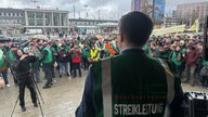 Sven Schmitte spricht als Streikführer der GDL auf einer Bühne. Er ist von hinten zu sehen, im Hintergrund stehen die Streikenden vor der Bühne.