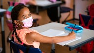 Eine Schülerin einer Grundschule sitzt mit Maske an ihrem Platz und macht Schulaufgaben.
