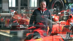 Reiner Ferling, Vorsitzender des Michael und Mick Schumacher Fan-Club Kerpen, steht zwischen Rennwagen, die Michael Schumacher gefahren hat