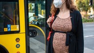 Eine Schwangere steht mit Maske neben einem Bus.