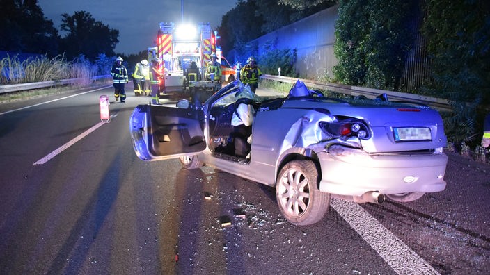 Das zerstörte Fahrzeug steht nach dem Unfall auf der Autobahn A57, Einsatzkräfte von Notarzt und Feuerwehr sind am Unfallort.