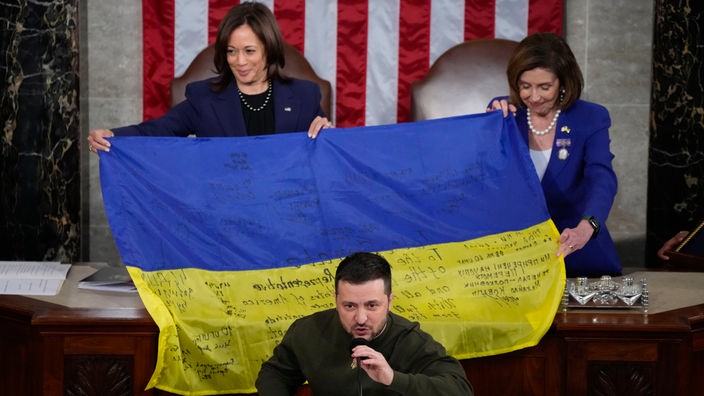 Wolodymyr Selenskyj, Präsident der Ukraine, spricht vor einer ukrainischen Flagge, die von Kamala Harris (l) und Nancy Pelosi gehalten wird