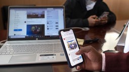 Ein Handybenutzer ist auf einer sozialen Plattform unterwegs. Auch im Hintergrund ist ein Computerbildschirm zu sehen auf welchem ein soziales Netzwekr geöffnet ist.
