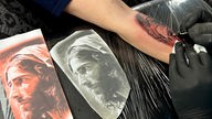 Zwei Vorlagen eines Jesus Tattoo-Motivs und ein Unterarm, der mit dem Tattoo bestochen wird. 