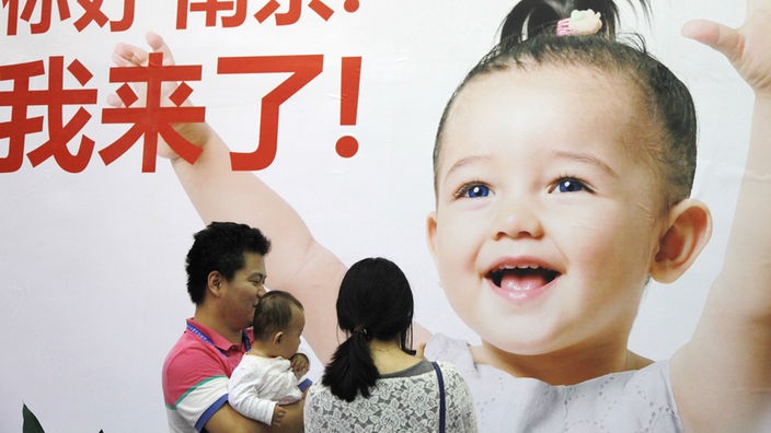 Eine dreiköpfige Familie steht vor einer Plakatwand, auf der ein kleines Mädchen zu sehen ist