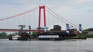 Das U-Boot, befestigt auf einem Transporterschiff, auf dem Niederrhein bei Kleve. Im Hintergrund ist die Rheinbrücke Emmerich zu sehen. 