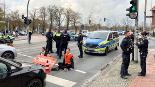 Aktivisten sitzen auf der Straße, umgeben von Polizeibeamten und Einsatzwagen. 