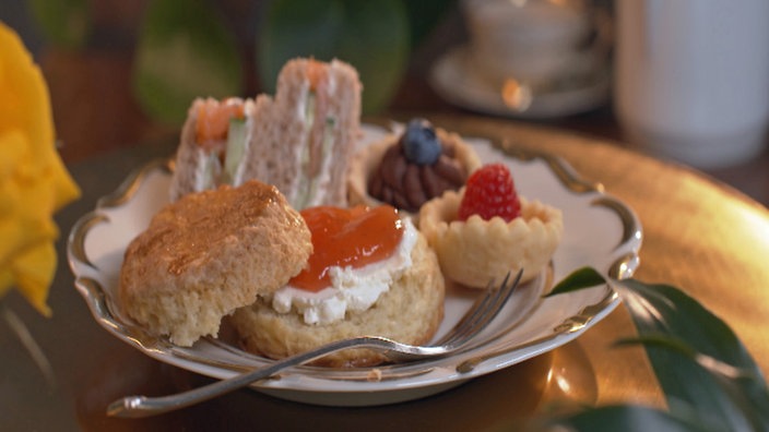 Mit Creme und Konfitüre beschmierte Scones, mit Lachs und Gurke belegte Sandwiches und mit Beeren verzierte Tartelettes auf einem altmodischen Kuchenteller mit einer silbernen Gabel.