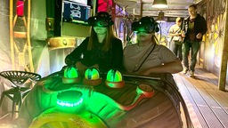 Zwei junge Frauen sitzen in einer Gondel und haben eine VR-Brille auf.