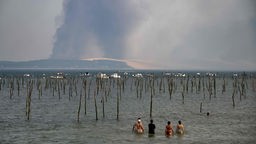 Eine Gruppe Menschen steht im Wasser und betrachtet dunkle Rauchwolken über der Pilat-Düne von Cap Ferret, die durch einen Waldbrand in der Nähe von La Teste im Südwesten Frankreichs entstanden sind. 