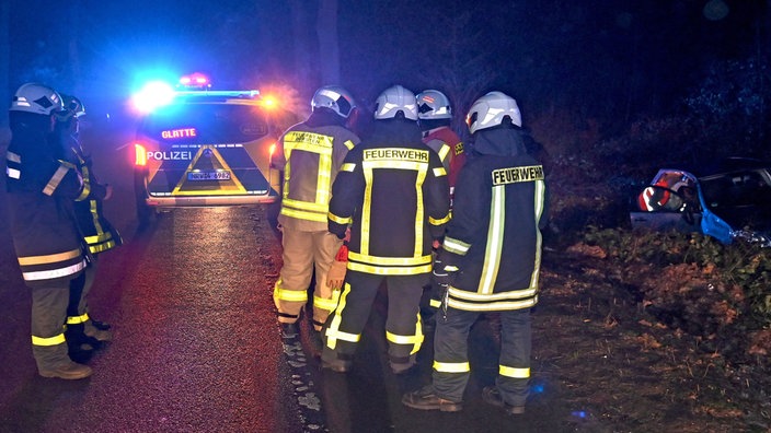 Im Einsatzfahrzeug der Polizei leuchtet auf einem Display der Hinweis "Glätte" an einer Unfallstelle. Einsatzkräfte der Feuerwehr stehen auf der Bundesstraße 58 an der Unfallstelle mit einem verunfallten Pkw