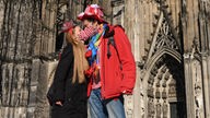 Ein kostümiertes Paar küsst sich vor dem Kölner Dom. Beide tragen Masken.
