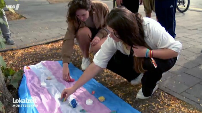Zwei junge Frauen legen Gedenksteine auf eine blau-rosa-weiß gestreifte Flagge, die auf dem Boden vor einem Baum liegt. 