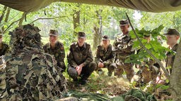 Auf dem Foto sind junge Menschen in Camouflage-Uniform der Bundeswehr, die einer Person im Tarnanzug zuhören.