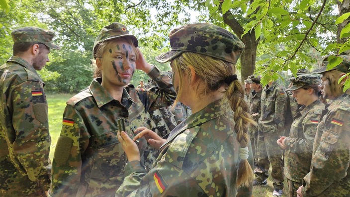 Auf dem Foto sind zwei blonde junge Frauen im Bundeswehr-Anzug. Die eine malt der anderen schwarze Streifen ins Gesicht.