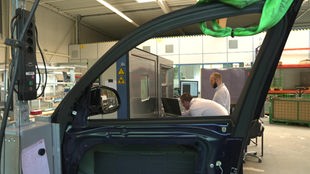 In einer Versuchsanlage wird eine Autortür mit einem Bremssystem geprüft. Im Hintergrund stehen zwei Forscher und prüfen die Ergebnisse an einem Computer.