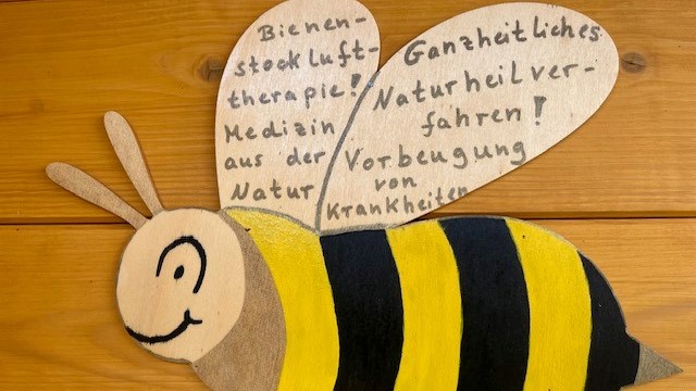 Eine gefilzte Bienen hängt an einer Holzwand, in ihren Flügeln stehen Erklärungen, worum es sich bei der Bienenstocklufttherapie handelt geschrieben