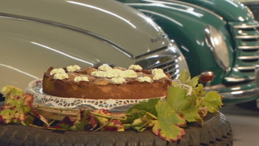 Der herbstlicher Birne-Helene-Kuchen steht auf einem Tisch. Im Hintergrund sind die Motorhauben von zwei Oldtimern zu sehen