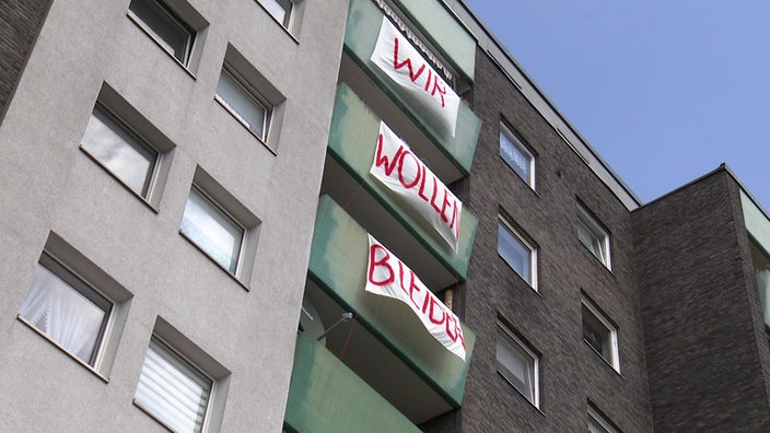 Auf dem Foto ist ein Haus mit grünen Balkonen, an dem drei Banner angebracht sind, die zusammen die Schrift "Wir wollen bleiben" ergeben.