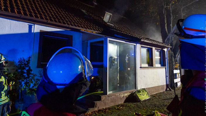 Zwei Feuerwehrmänner stehen vor einem rauchenden Einfamilienhaus