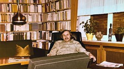 Der Sammler Bruno Schröder sitzt auf einem Sessel vor einem hohen Bücherregal und schaut in die Kamera.