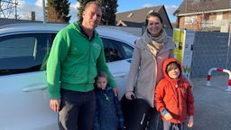 Eine Familie, beide Elternteile und zwei kleine Kinder, vor dem Nachbarschaftsauto stehend
