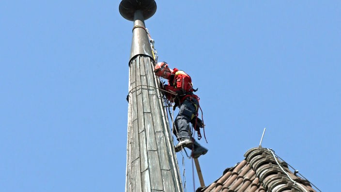 Ein Mann mit Helm klettert durch Seile gesichert eine Kirchturmspitze hoch
