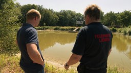 Zwei Männer von der Feuerwehr stehen vor einem See.