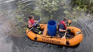 Zwei Mitarbeitende der Feuerwehr in einem Schlauchboot auf der Hessel, mit Kescher und Paddel in der Hand