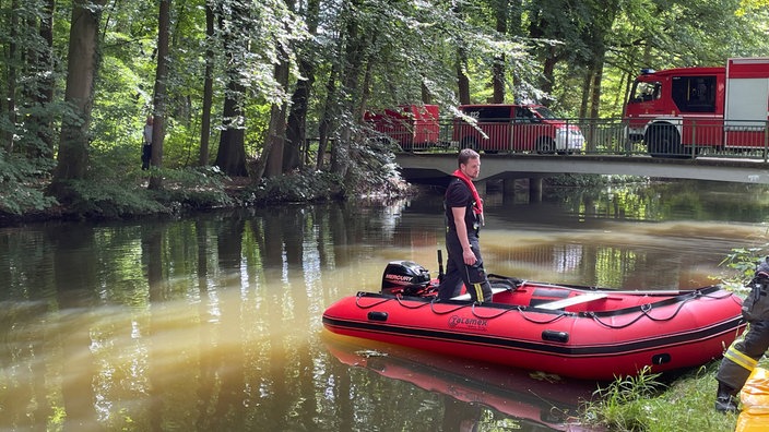 Ein Schlauchboot am Ufer der Hessel, darauf steht ein Mann, im Hintergrund eine Brücke auf der Feuerwehr-Fahrzeuge stehen