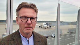 Ein Mann mit Brille steht vor einer Scheibe, dahinter ein Flugzeug