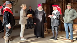 Eine Frau hält ein Brot und ist umgeben von Männern in Trachten. 