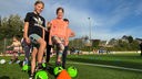 Zwei Mädchen in Sportkleidung stehen auf einem Fußballplatz. Ihre rechten Beine haben sie auf einen Fußball gestützt.